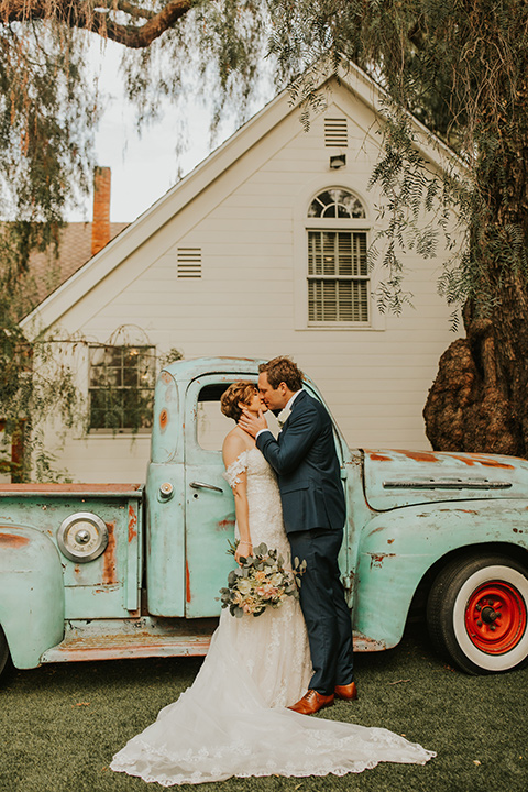  a romantic rustic garden wedding with the groom in a blue suit and floral tie and the bride in a lace gown - couple kissing in front of rustic truck 
