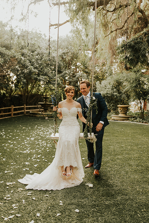  a romantic rustic garden wedding with the groom in a blue suit and floral tie and the bride in a lace gown - groom pushing bride on the swing 