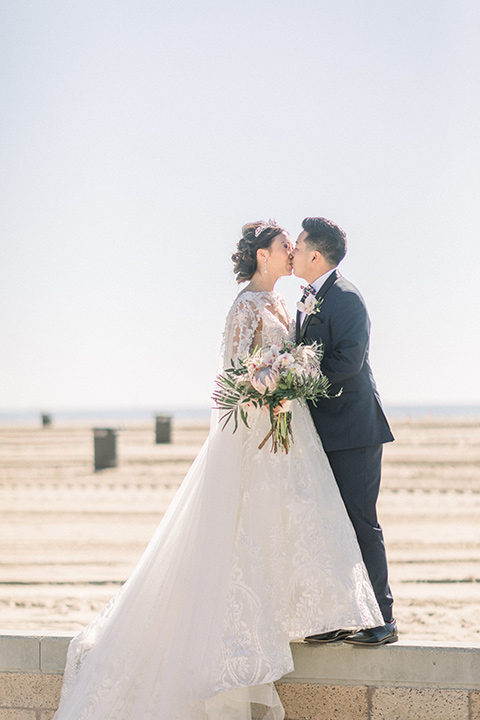  a grand cultural wedding with lion dancers and gold details – bride in a long gown with cape and groom in navy shawl tuxedo – couple by the ocean 