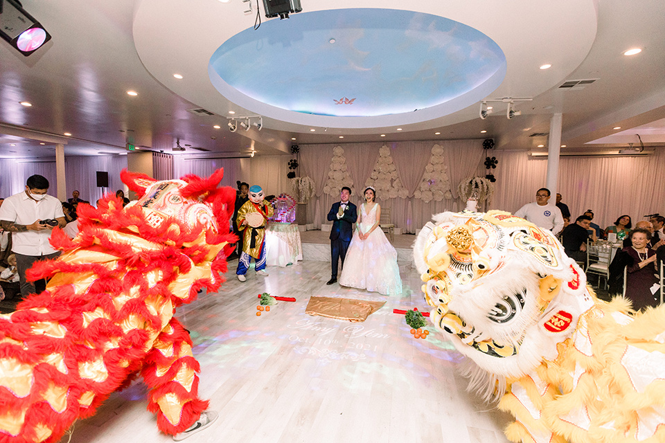  a grand cultural wedding with lion dancers and gold details – bride in a long gown with cape and groom in navy shawl tuxedo – lion dancers 