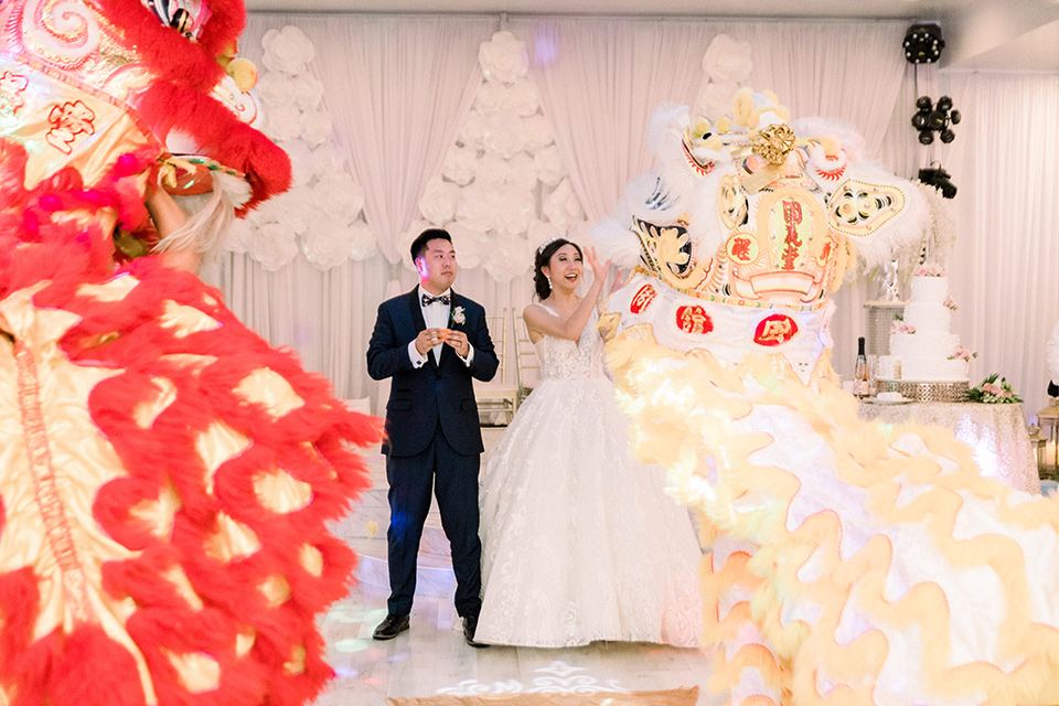  a grand cultural wedding with lion dancers and gold details – bride in a long gown with cape and groom in navy shawl tuxedo – lion dancers 