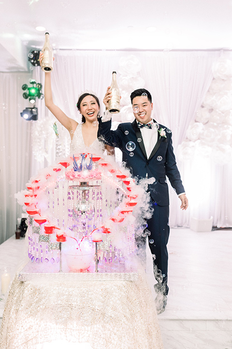  a grand cultural wedding with lion dancers and gold details – bride in a long gown with cape and groom in navy shawl tuxedo – couple pouring drinks 