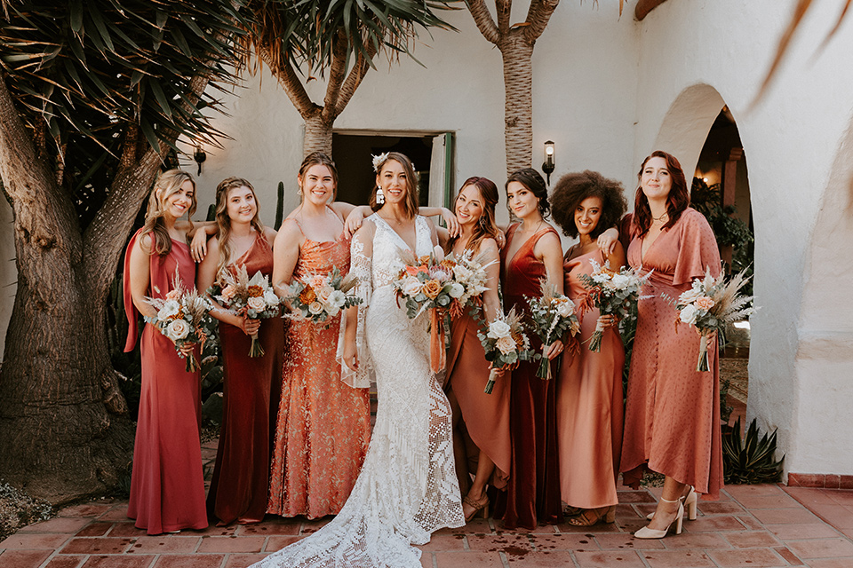  boho beach wedding with Spanish inspired architecture with the bride in a lace gown and the groom in a black tuxedo – bridesmaids in orange-toned dresses