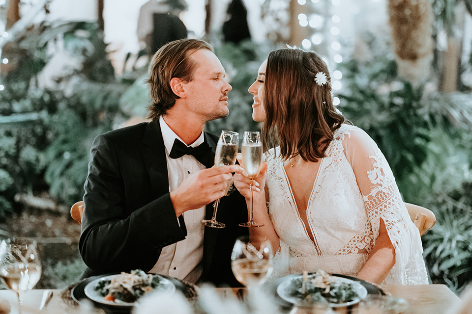  boho beach wedding with Spanish inspired architecture with the bride in a lace gown and the groom in a black tuxedo – couple cheersing