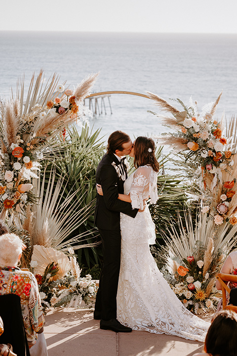  boho beach wedding with Spanish inspired architecture with the bride in a lace gown and the groom in a black tuxedo – first kiss