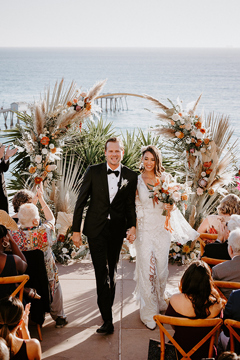  boho beach wedding with Spanish inspired architecture with the bride in a lace gown and the groom in a black tuxedo – walking down the aisle 
