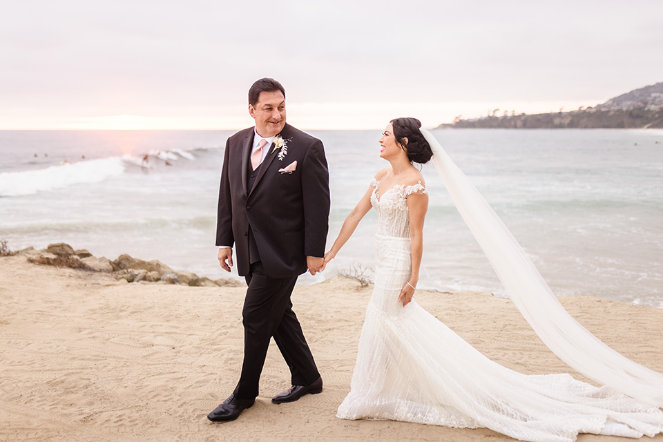  glitz and glam wedding by the beach with the bride in a lace crystal gown and the groom in a black tuxedo – couple walking on the sand