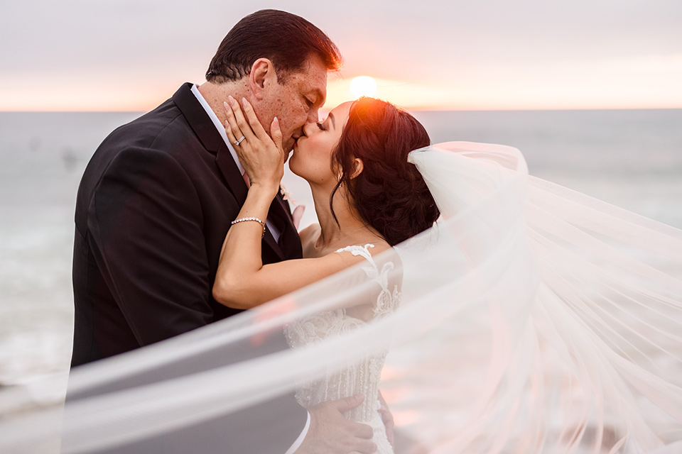  glitz and glam wedding by the beach with the bride in a lace crystal gown and the groom in a black tuxedo – kissing on the sand