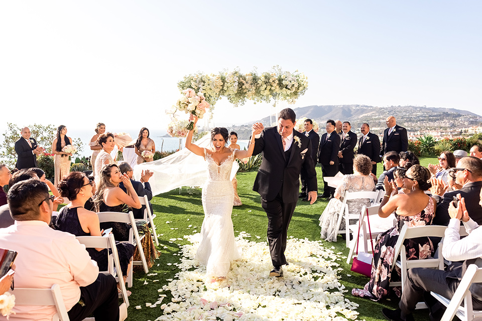  glitz and glam wedding by the beach with the bride in a lace crystal gown and the groom in a black tuxedo – walking down the aisle