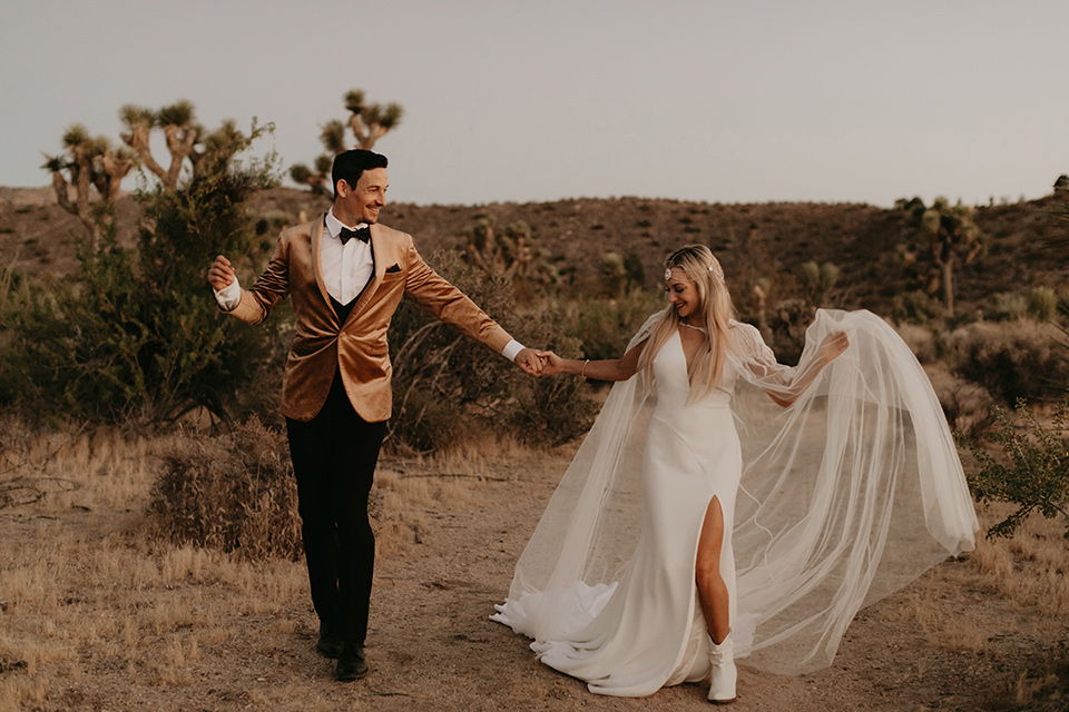  joshua tree wedding with the groom in a gold velvet tuxedo and a black tuxedo and the bride in a flowing gown with bridal wings – running in the desert