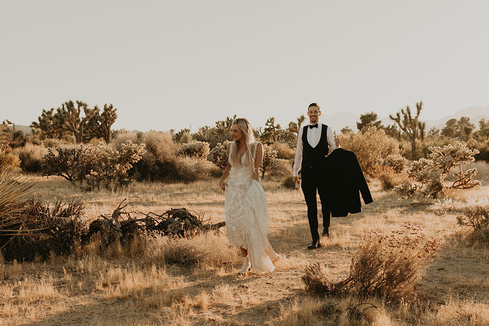  joshua tree wedding with the groom in a gold velvet tuxedo and a black tuxedo and the bride in a flowing gown with bridal wings – couple walking in the desert