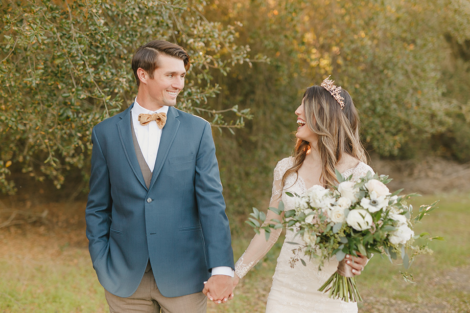  fairytale wedding in the meadow with the bride in a lace long sleeve gown and the groom in a slate blue and café brown look – bride and groom holding hands 