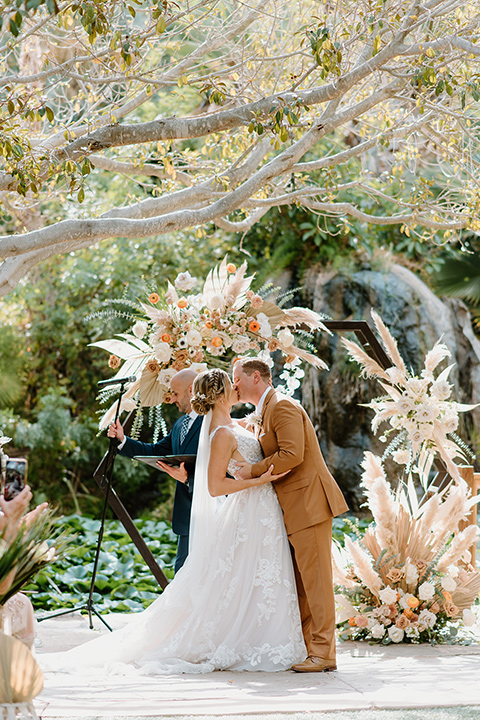  earth-toned boho wedding with the bridesmaids in green dresses, groomsmen in tan suits, and the groom in a caramel suit – couple first kiss 