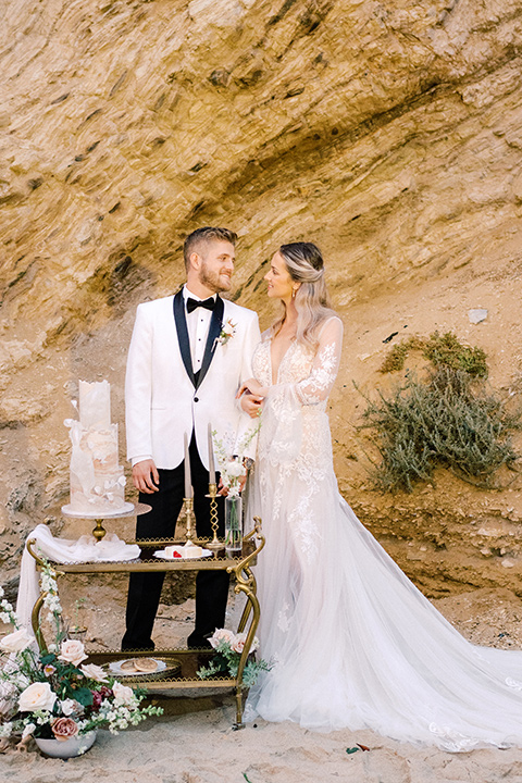  beach side wedding with the bride in a flowing lace gown and the groom in a white tuxedo – couple with cake 