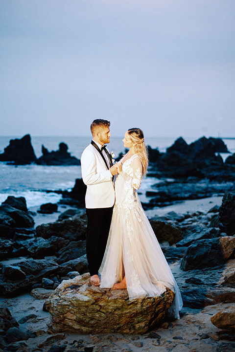  beach side wedding with the bride in a flowing lace gown and the groom in a white tuxedo – couple by the water