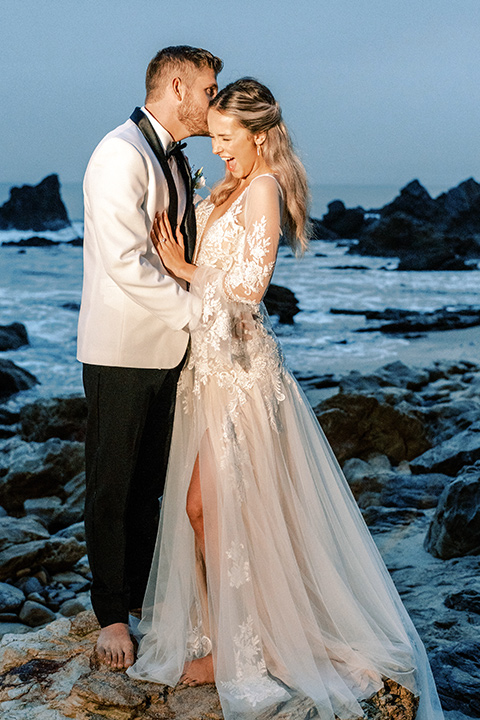  beach side wedding with the bride in a flowing lace gown and the groom in a white tuxedo – couple by the water 