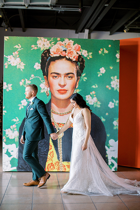  museum of latin american art wedding with 2 different styles – one bride in a fitted gown and the groom in a grey suit and the other bride in a lace gown and the groom in a green suit – couple walking by mural 