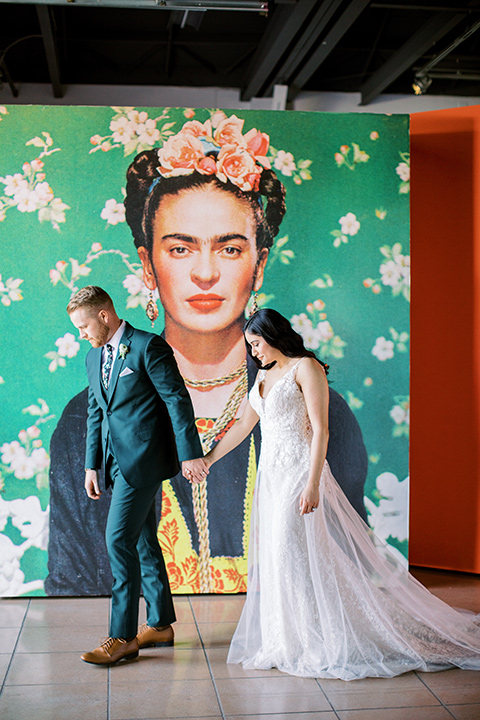  museum of latin american art wedding with 2 different styles – one bride in a fitted gown and the groom in a grey suit and the other bride in a lace gown and the groom in a green suit – couple walking by mural 