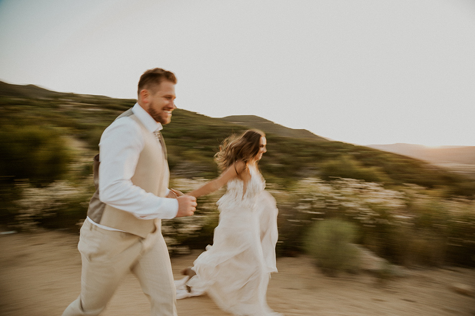  wildflower wedding in a field with the bride in a flowing gown and bridal wings, and the groom in a tan suit and bolo tie – couple running