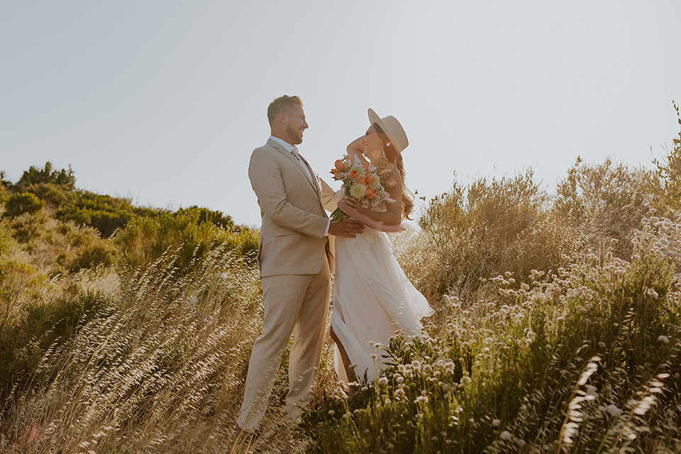  wildflower wedding in a field with the bride in a flowing gown and bridal wings, and the groom in a tan suit and bolo tie – couple laughing