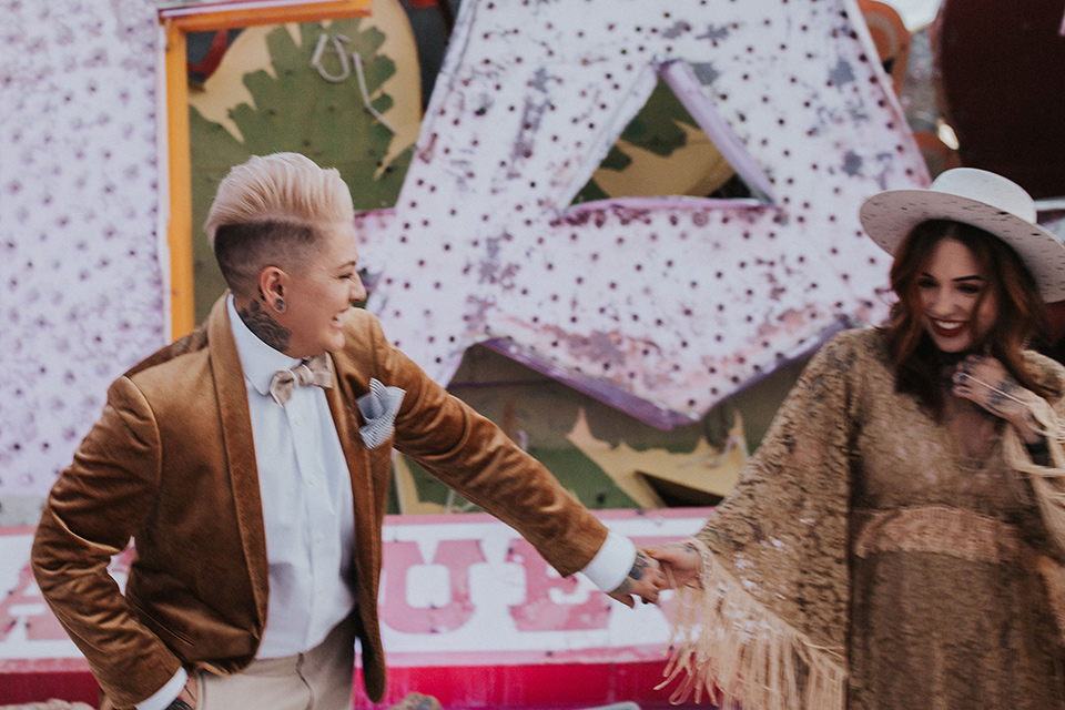  same sex wedding with two brides at the neon museum in las vegas with one brief is a bohemian ballgown and the other in a gold velvet tuxedo coat and tan pants – couple walking 