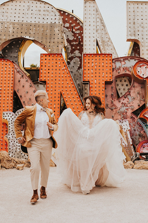  same sex wedding with two brides at the neon museum in las vegas with one brief is a bohemian ballgown and the other in a gold velvet tuxedo coat and tan pants – couple walking 