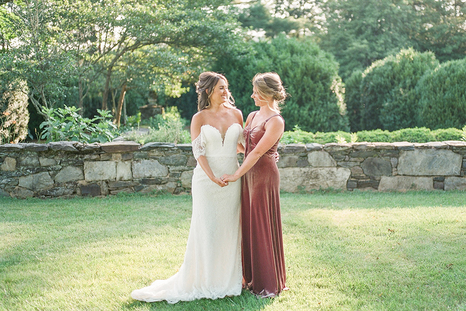  blush and burgundy wedding in the woods with the bride in a boho lace gown, the groom in a burgundy shawl tuxedo, and the bridesmaids in a dusty blush velvet gown – bride and bridesmaid
