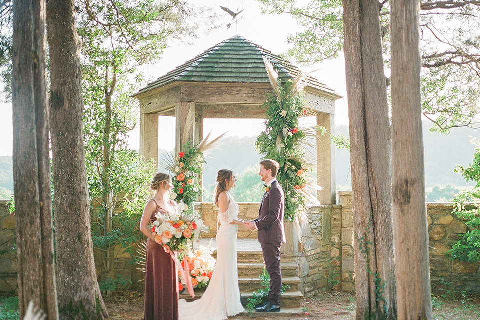  blush and burgundy wedding in the woods with the bride in a boho lace gown, the groom in a burgundy shawl tuxedo, and the bridesmaids in a dusty blush velvet gown – ceremony