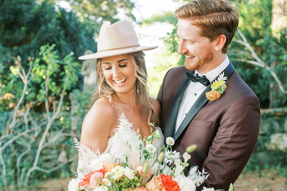  blush and burgundy wedding in the woods with the bride in a boho lace gown, the groom in a burgundy shawl tuxedo, and the bridesmaids in a dusty blush velvet gown – couple laughing
