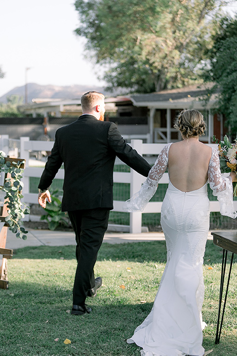  a lavender and black wedding in a garden – with the bride in a long sleeve lace gown and the groom in a black tuxedo – couple walking down the aisle 