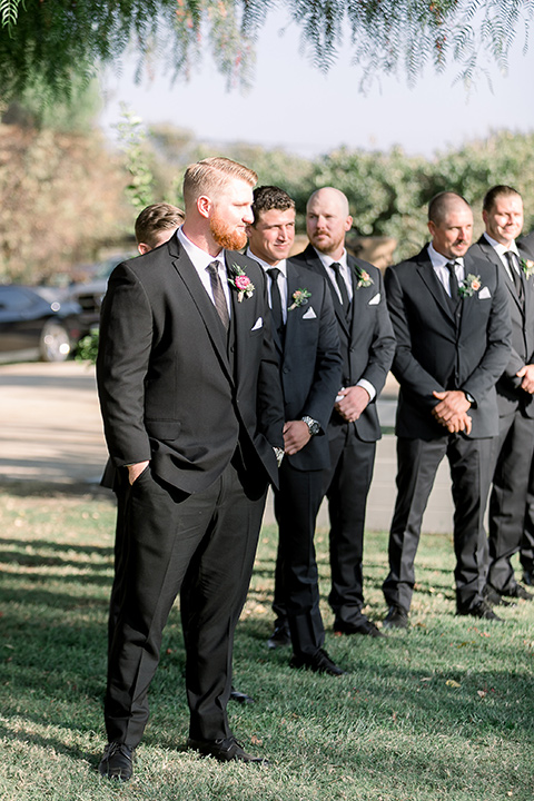  a lavender and black wedding in a garden – with the bride in a long sleeve lace gown and the groom in a black tuxedo – groom watching the bride walk down the aisle 