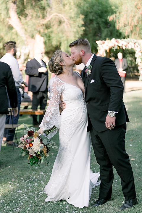  a lavender and black wedding in a garden – with the bride in a long sleeve lace gown and the groom in a black tuxedo – couple kissing down the aisle