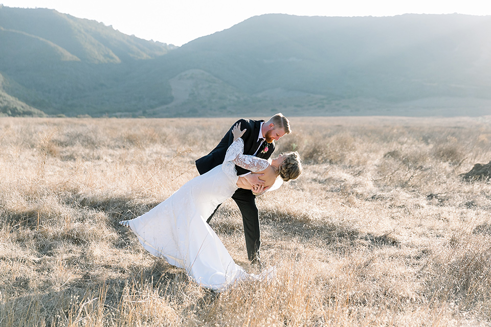  a lavender and black wedding in a garden – with the bride in a long sleeve lace gown and the groom in a black tuxedo – kissing in the meadow