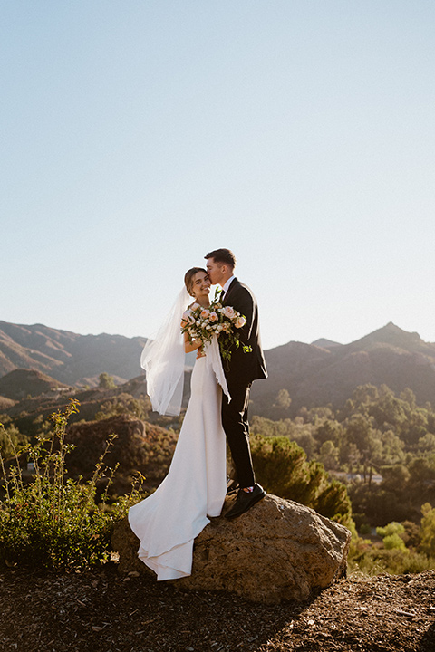  blush and beige wedding with a chic modern twist – the bride in a cap sleeved fitted gown and the groom in a black suit – couple on cliff 
