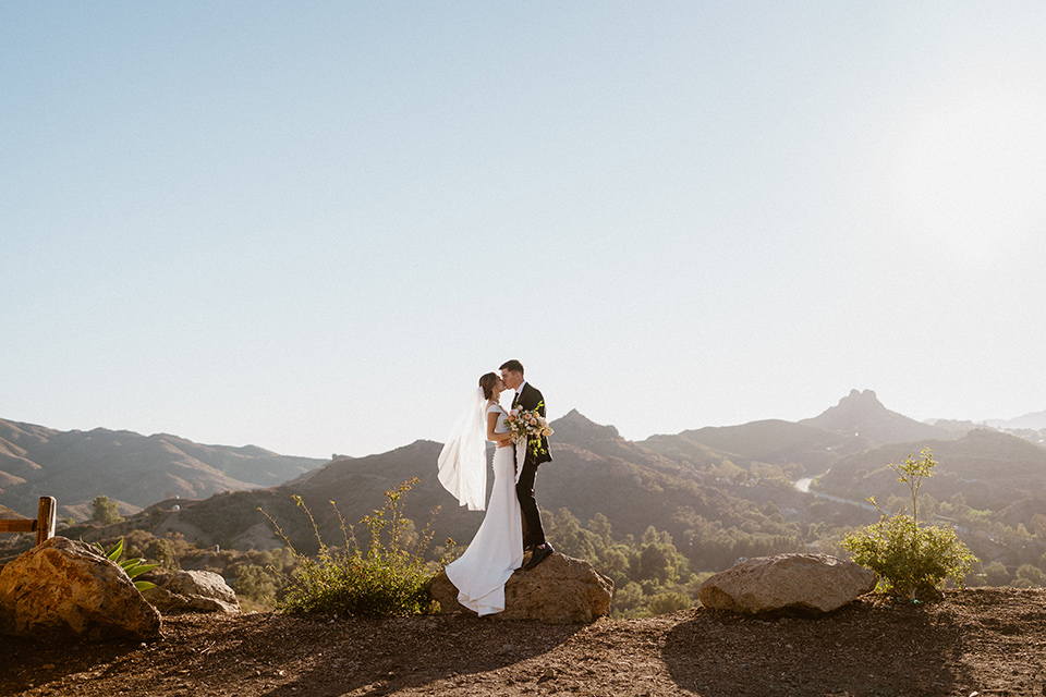  blush and beige wedding with a chic modern twist – the bride in a cap sleeved fitted gown and the groom in a black suit – couple on cliff