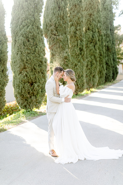  English garden romantic wedding with the bride in an off-the-shoulder gown and the groom in a tan suit – bride and groom kissing by the hedges 