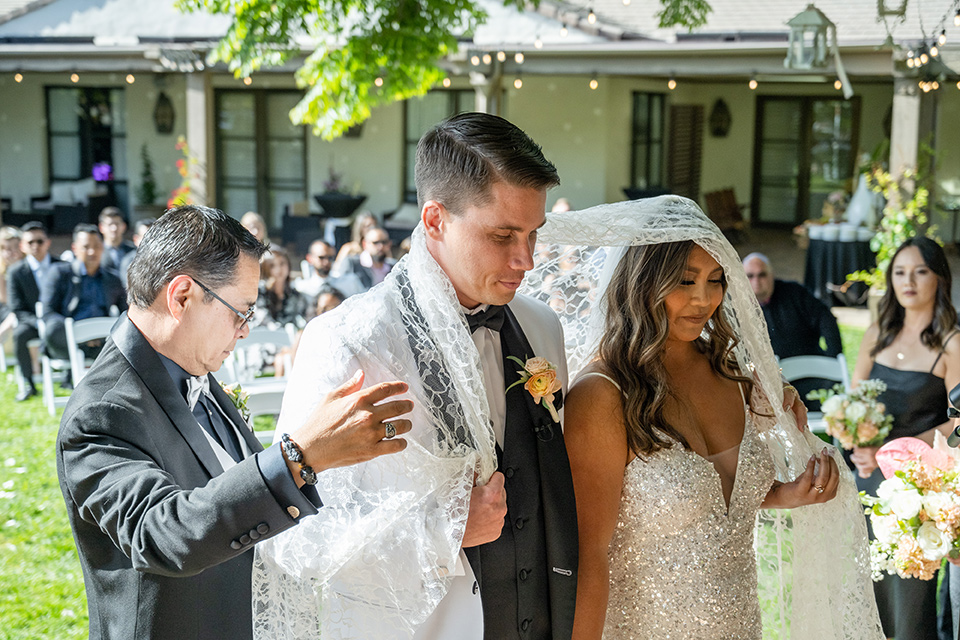  a modern black and white wedding with crystal details -the bride in a lace formfitting gown and the groom in a white + black tuxedo – couple at the ceremony