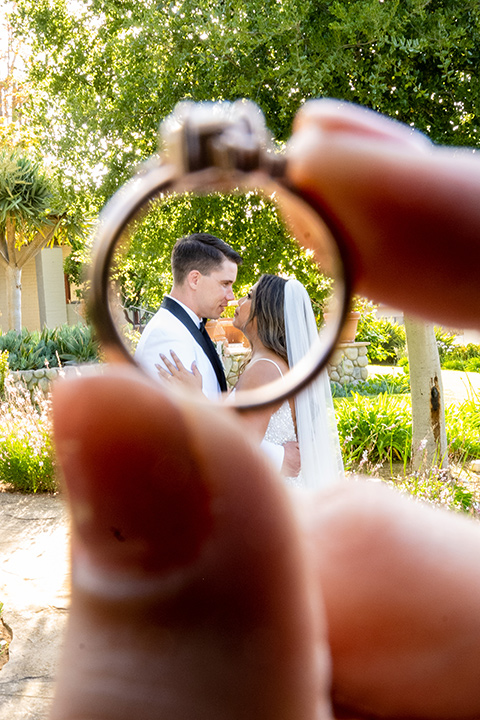  a modern black and white wedding with crystal details -the bride in a lace formfitting gown and the groom in a white + black tuxedo – looking at the couple through their wedding rings 