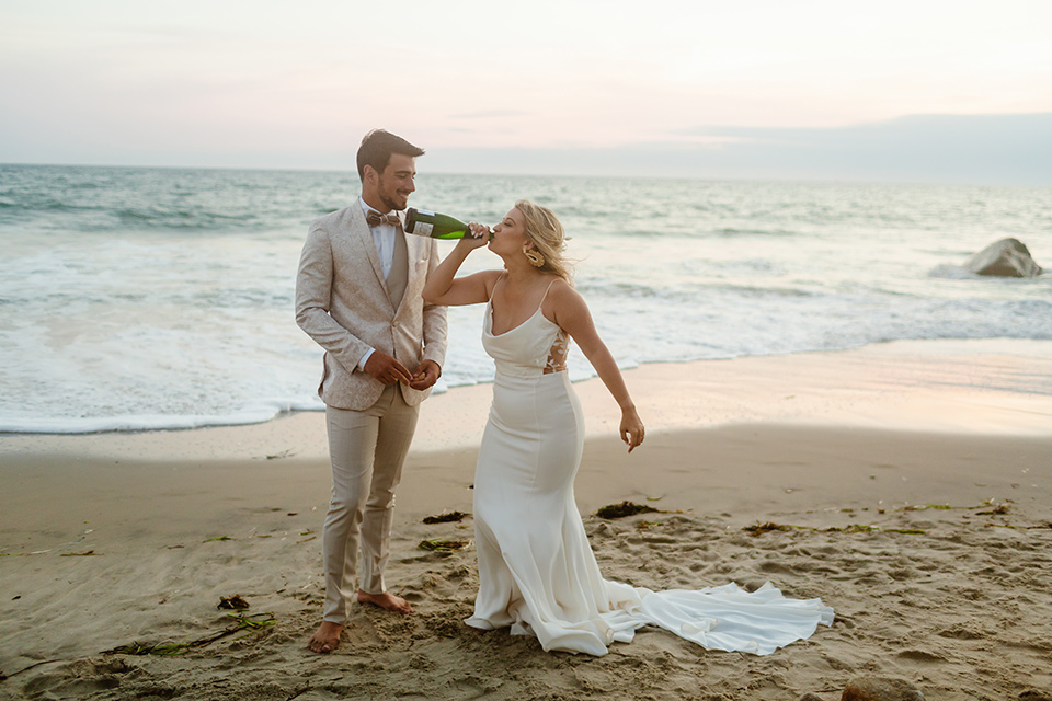  a neutral beachy elopement with the groom in a tan suit and the bride in a white lace dress – drinking champagne