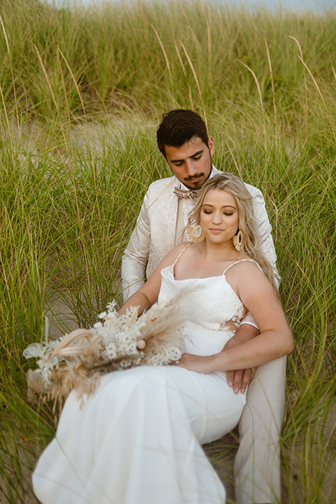  a neutral beachy elopement with the groom in a tan suit and the bride in a white lace dress – sitting in the grasses 