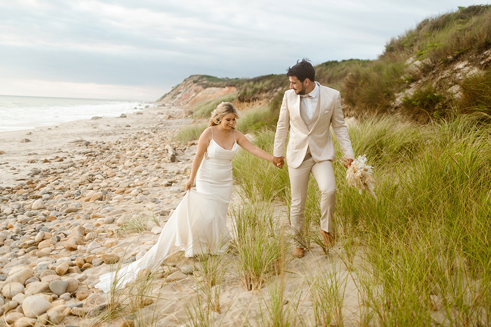  a neutral beachy elopement with the groom in a tan suit and the bride in a white lace dress – couple walking holding hands 